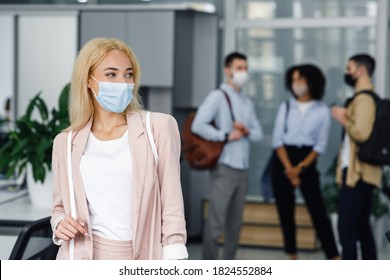 Return To Work After Lockdown And New Normal. Woman In Protective Mask In Business Suit Looks To The Side In Corridor Of Modern Office With Blurred Colleagues During Covid-19, Free Space