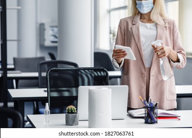 Return To Work, Workplace Hygiene And New Normal. Blond Woman In Business Suit In Protective Mask With Paper Towel And Spray Disinfect Table In Modern Office Interior During Covid-19, Cropped