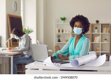 Return To Workplace After Coronavirus Pandemic Lockdown. Portrait Of Black Woman In Face Mask Working On Laptop In Studio Office. Architect Or Designer Using Computer Sitting At Desk With Paper Rolls