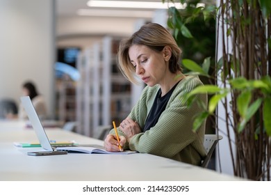 Returning To Study As Mature Student. Focused Middle-aged Female University Professor Taking Notes In Day Planner While Working On Laptop In Empty Library, Selective Focus. Academic Work Concept