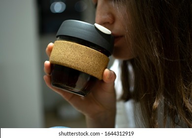 Reusable Coffee Eco Cup. Toughened Glass Cup & Natural Cork Band. Secure Silicon Lid. Friendly, Spill Proof Travel Mug With Lid | Black.A Young Girl Drinks From A Reusable Cup. Keep Cup 