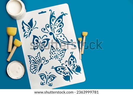 Reusable flexible plastic stencil for graphical design, white paint and round sponge foam brushes on a blue background