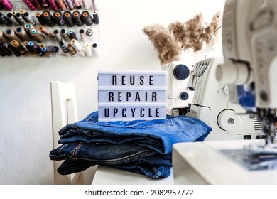 Reuse, Repair, Upcycle Text On Light Board On Sewing Machines Background. Stack Of Old Jeans, Denim Clothes, Scissors, Thread And Sewing Tools In Sewing Studio. Denim Upcycling Ideas, Using Old Jeans