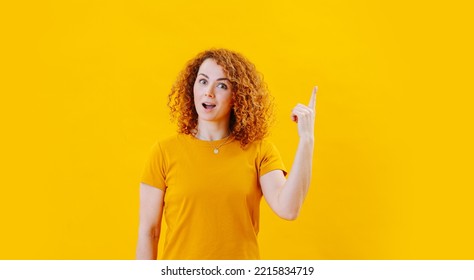 Revelation Face Of A Young Woman With Curly Ginger Hair Pointing Up. Over Yellow Background. Wearing An Orange Shirt.