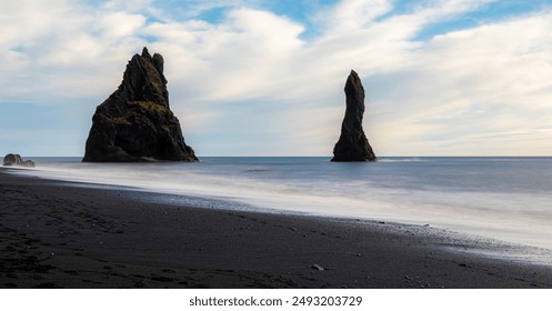 Reynisdrangar spires at Reynisfjara Black Sand Beach, Iceland. A rocky beach with large rocks in the foreground. The sky is cloudy, and the water is calm. Scene is serene and peaceful - Powered by Shutterstock