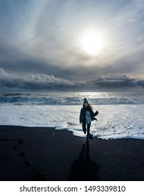 Reynisfjara, Iceland - October 16,2018: Smiling Woman Running From The Foamy Ocean Towards The Camera On The Black Sand Beach.  We Can Also See The Bright Sun In The Back As Well As The Clouds. 