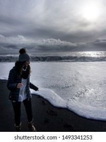 Reynisfjara, Iceland - October 16,2018: Smiling Woman Running From The Foamy Ocean Towards The Camera On The Black Sand Beach.  We Can Also See The Bright Sun In The Back As Well As The Clouds. 