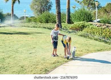 Rhodes, Greece - July 07 2017: Brothers In Soaking Wet Clothes Play With Watering System Near Green Field For Sports Activities. Preschooler And Schoolboy Have Fun On Sunny Day