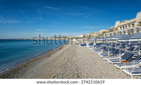 A rhythmic pattern of blue and white striped sun umbrellas and lounge chairs on a pebble beach overlooking the turquoise Mediterranean Sea in Nice, France 