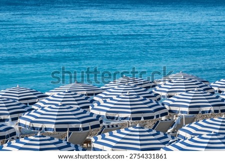A rhythmic pattern of blue and white striped sun umbrellas and lounge chairs on a pebble beach overlooking the turquoise Mediterranean Sea in Nice, France 
