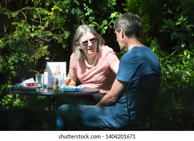 Ribchester, Lancashire/UK - May 14th 2019: Caucasian Middle Age Couple Sat Talking At A Garden Café Bistro Table In The Sunlight With Shaded Green Bushes In The Background