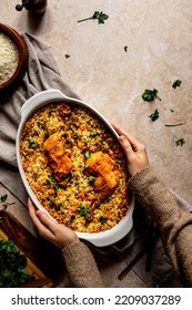 Rice Casserole Dish With Vegetables And Chicken Drumsticks In A Rustic Set Up Top View With Female Hands Holding The Oval Dish On A Brown Table