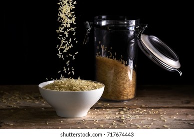 rice grains falling into a white bowl beside a glass jar with rice on a rustic wooden table against a dark background, food concept against hunger and for world nutrition, selected focus - Powered by Shutterstock