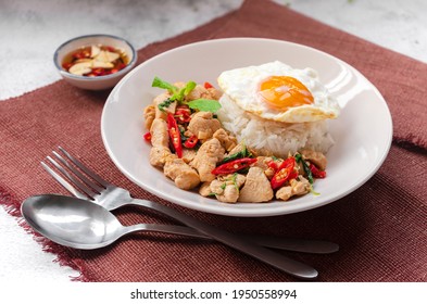 Rice topped stir-fried basil with chiken and a fried egg on white plate serving on brown table cloth with fish sauce - Thai street food