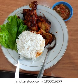 The Rice Is In A White Plate With Grilled Chicken, Green Lettuce Has A Spoon And A Fork On The Edge, You Can See Chili Sauce On A Small Blue Plate On A Wooden Table.