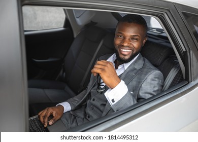 Rich african american young businessman smiling through open car window. Cheerful handsome black man in stylish suit going to business trip by car, sitting on back seat and looking through window