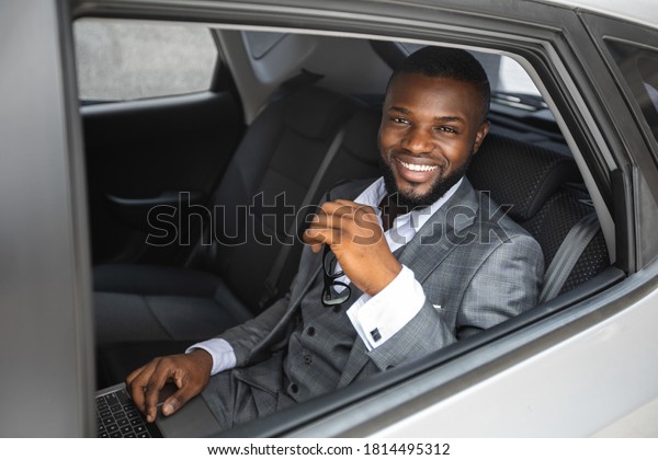 Rich african american young businessman smiling\
through open car window. Cheerful handsome black man in stylish\
suit going to business trip by car, sitting on back seat and\
looking through window