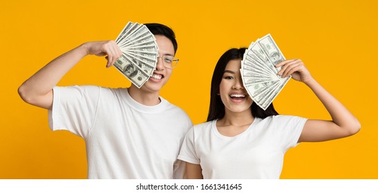 Rich Asian Couple Covering Faces With Bunch Of Money Over Yellow Studio Background, Panorama