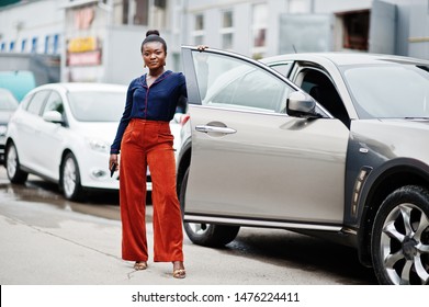 Rich Business African Woman In Orange Pants And Blue Shirt Posed Against Silver Suv Car With Opened Door.