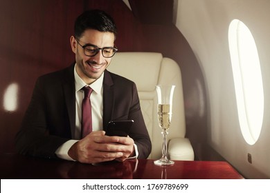 Rich business man sitting on board of private jet, drinking champagne and reading messages or checking business news on phone - Powered by Shutterstock