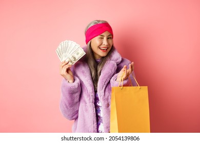 Rich And Fashionable Asian Senior Woman Wasting Money In Shops, Holding Shopping Bag And Dollars, Winking Happy At Camera, Pink Background