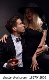 A Rich Handsome Man Drinks Whiskey With A Blonde Mistress In The Evening In Low Light. Photos Of A Couple In Love