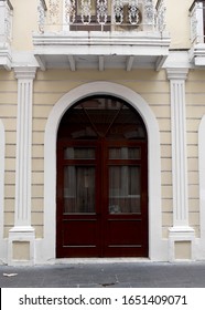 A Rich Mahogany Wood Rounded Door Set Into A Cream Colored Brick Wall Framed By A White Arch Below A Raft Iron Balcony.