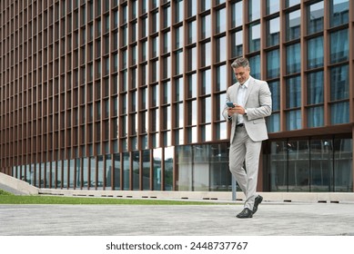 Rich middle aged businessman using cellphone walking on city street, busy older business man leader investor, mature male executive in suit holding mobile phone going near office building, full body. - Powered by Shutterstock