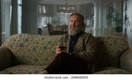 Rich Senior Man Enjoy Retirement Life Cognac In Luxury Home Interior. Confident Old Gentleman Drinking Whiskey Glass On Vintage Sofa. Intelligent Elderly Grandparent Relaxing Alone Alcohol Drink