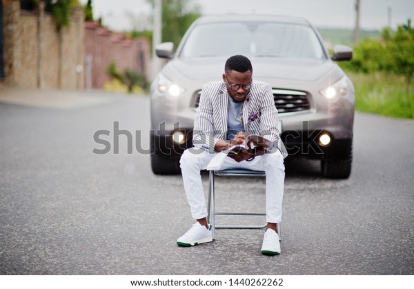 Rich and stylish african american man in blazer and\
white pants, eyeglasses with magazine sitting in chair against suv\
car.