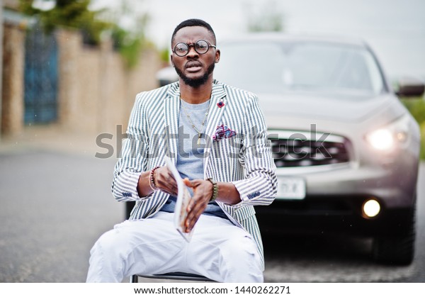 Rich and stylish african american man in blazer and\
white pants, eyeglasses with magazine sitting in chair against suv\
car.