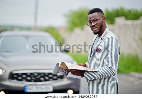 Rich and stylish african\
american man in blazer and eyeglasses read magazine against his suv\
car.