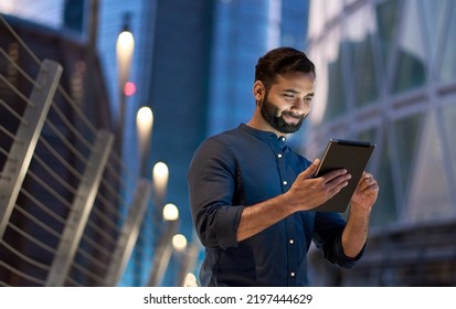 Rich successful eastern indian business man professional entrepreneur standing outdoors street holding using digital tablet online technology in night big city and urban lights   skyscrapers 