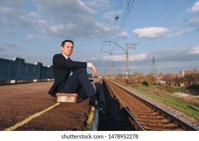 A rich upset and offended businessman is sitting and waiting for a train on the platform. Dismissal, depression and headache. Stylish man in a black suit on the background of clouds and sky.