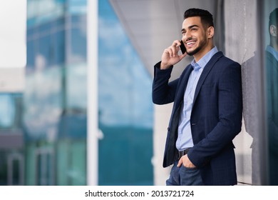 Rich young middle-eastern businessman having phone conversation next to modern office building, panorama with copy space. Happy arab entrepreneur making business call, using cellphone