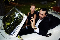 Richly Dressed Wealthy And Famous Couple Sitting Inside Cabriolet Luxury Car, Successful Man Sitting Behind Convertible Steering Wheel With Its Ok Hand Gesture, Happy Man And Woman Enjoying The Life
