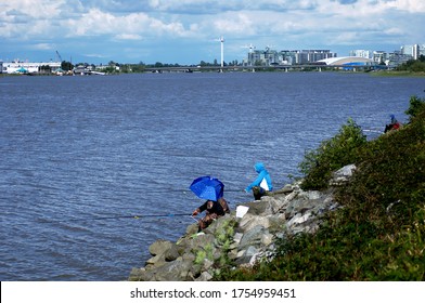 RICHMOND, BC,CANADA- 06/06/2020: Fishermen Along The Fraser River In Richmond, Keeping Out Of The Sun With An Umbrella, With Construction Cranes In The Background.