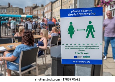 Richmond, North Yorkshire, UK - August 1, 2020: A Sign Asking People To Observe Social Distancing With People Sat Outside At A Restaurant Eating In The Background