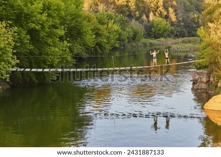 A rickety wooden suspension bridge spans across a river in the tranquil morning, and two young boys are seen making their way across it. The river flows peacefully beneath, reflecting the lush greener