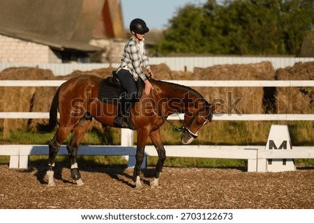 Rider on horse practices basic dressage exercises in open arena near stable, sitting in saddle and wearing helmet. Racing, horse care, and training