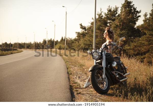 Riding a motorcycle. A beautiful girl is sitting\
on a motorcycle. Day. Background blue sky, forest, road. In short\
black shorts and a white T-shirt. Straightens her hair and looks at\
the sunset.