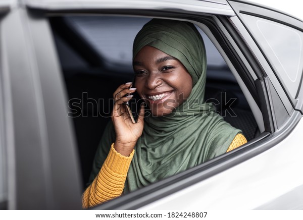 Riding\
Taxi. Cheerful Black Muslim Woman In Hijab Talking On Cellphone\
While Sitting On Backseat Of Car, Enjoying Mobile Communication,\
Looking Through Opened Window And Smiling,\
Closeup