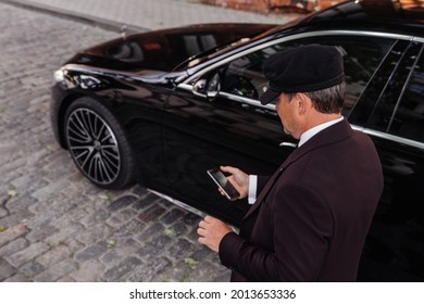 Riga, Latvia 21 July 2021 Mercedes Benz S500 AMG S Class W223 Brand New Model Stand On Parking Lot, Limousine Transfer Chauffeur With Chauffeur Hat, Stands By Luxury Car. 
Holding Mobile Phone In Hand