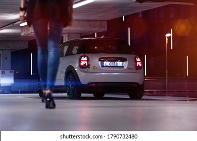 Riga, Latvia 6 August 2020 MINI Electric Cooper SE Stand At Parking Lot In Garage At Night Time.Led Lights On. White With Yellow Details. Countryside Panoramic View. Eco Life Woman Is Walking To Car