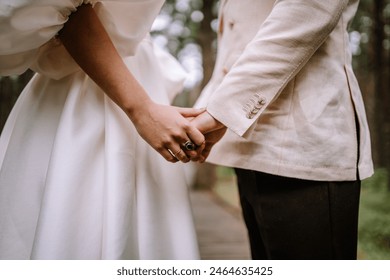 Riga, Latvia, - August 26, 2024 - Close-up of a bride and groom holding hands, showcasing the groom's suit and the bride's dress in a forest. - Powered by Shutterstock