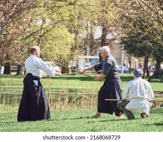 Riga, Latvia - May 8, 2022: Two aikido fighters training with stick in the public park. Pupil sitting and watching. Blooming sakura at the background.