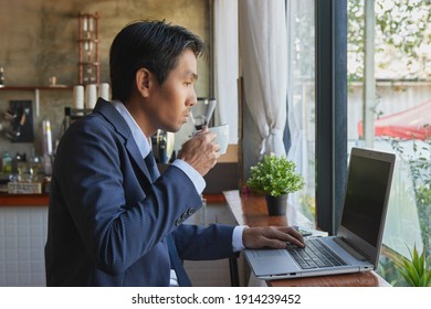 Der richtige asiatische Geschäftsmann in Suit und Tie Hold Coffee Cup und Tippen Laptop in Coffee Shop Scene. Asiatischer Geschäftsmann arbeitet von überall aus mit Technologie