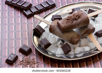 Right Chocolate Ice Cream On A Stick On A Silver Platter With Ice And Pieces Of Chocolate Near Chocolate Pieces Left Empty Space On A Chocolate Background. Chocolate Ice Cream Popsicles. Horizontal.