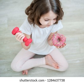 Right Choice. Little Cute Girl Holding Dumbbell In One Hand And Pink High-calorie Donut In Another. Kid Trying To Make Decision Between Healthy And Unhealthy Food. Dieting, Sport, Lifestyle. Top View