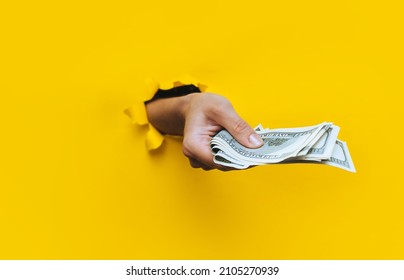 A right female hand appears in the hole in torn yellow paper and squeezes several hundred-dollar bills. Concept of poverty, benefits, scholarships, and stinginess. Isolated. Copy space.